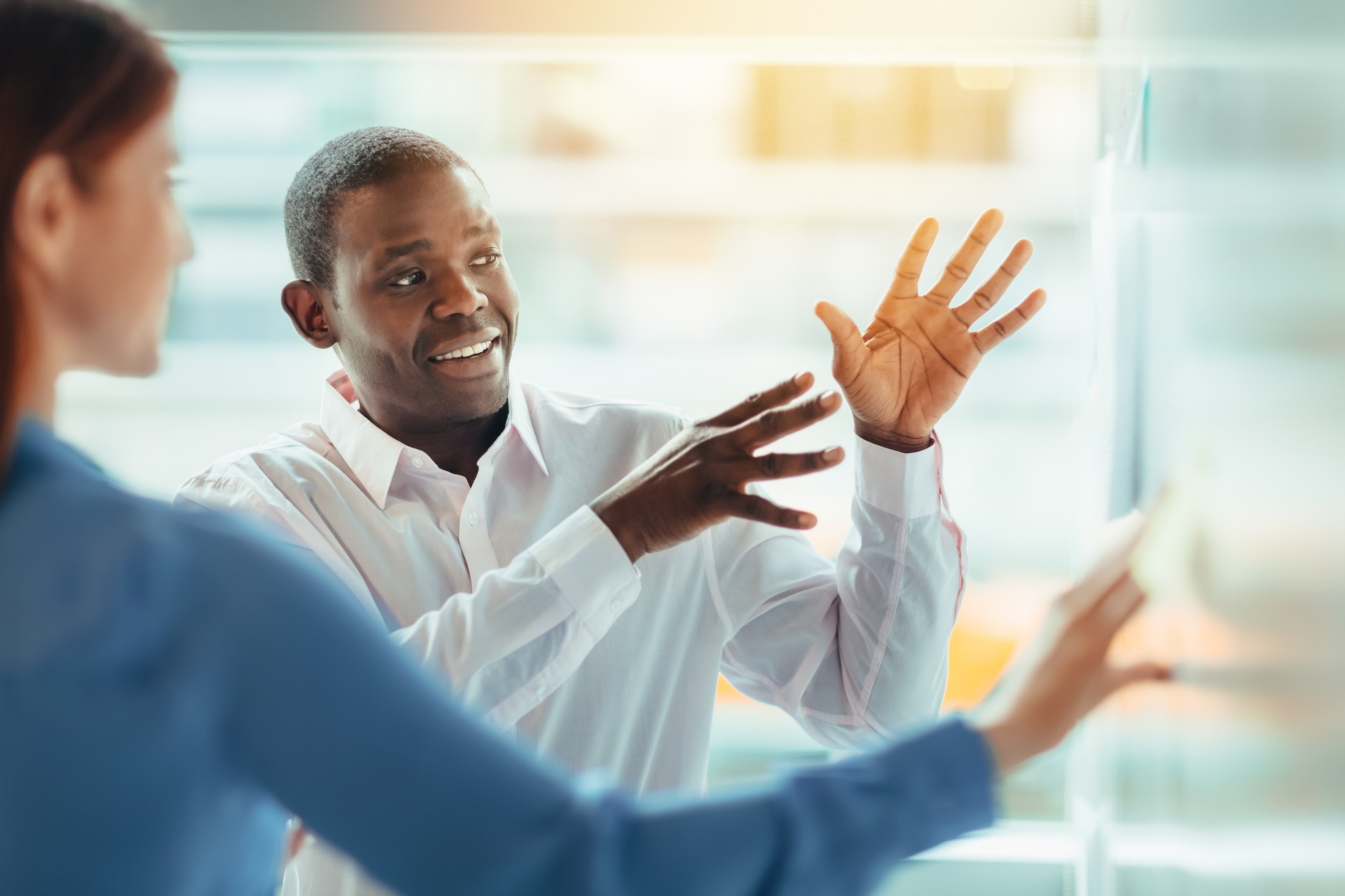 Side view of multiracial group of business persons work colleagues with Black mid adult man collaborating brainstorming sharing ideas pointing hand gesture to notes diagram on whiteboard visual aid standing in office meeting together with window