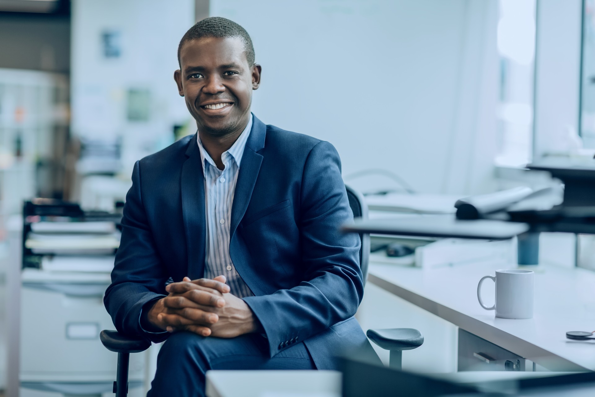 Portrait of Black mid adult businessman student sitting smiling happy positive emotion with hands clasped in bright business office in suit